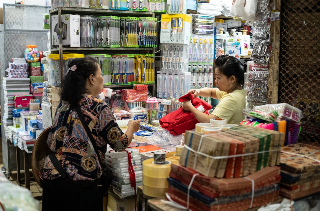 Women interacting in a bustling Dong Xuan Market shop filled with household goods in Hanoi, Vietnam.