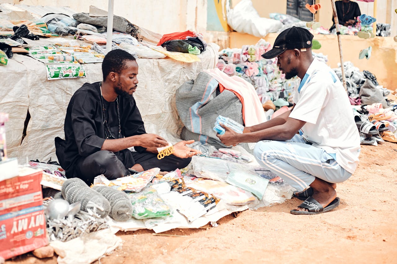 Two men engaging in a marketplace deal outdoors with various goods on display.
